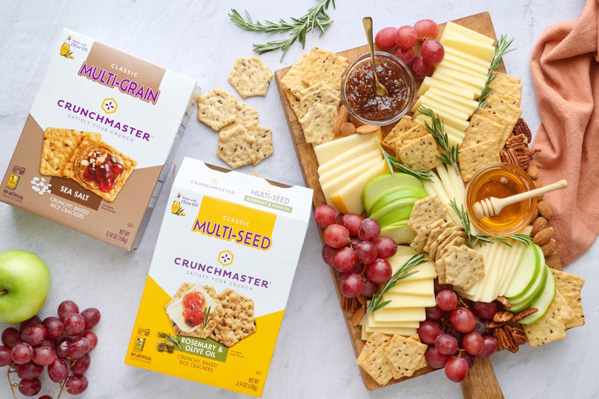 Fruit and Cheese platter featuring Crunchmaster Rosemary & Olive Oil Crackers and Crunchmaster Sea Salt Crackers, with cheese slices, grapes, apple slices, mixed nuts, jam, and honey.