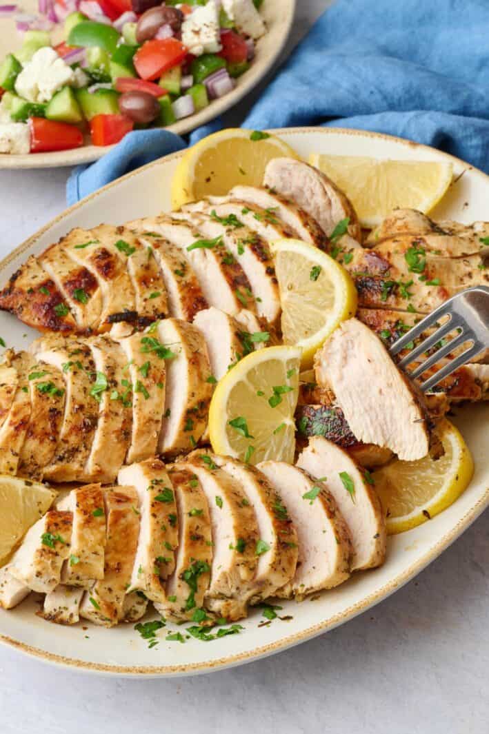 Sliced Greek chicken breasts on a platter with a fork lifting up a bite to show juicy texture.