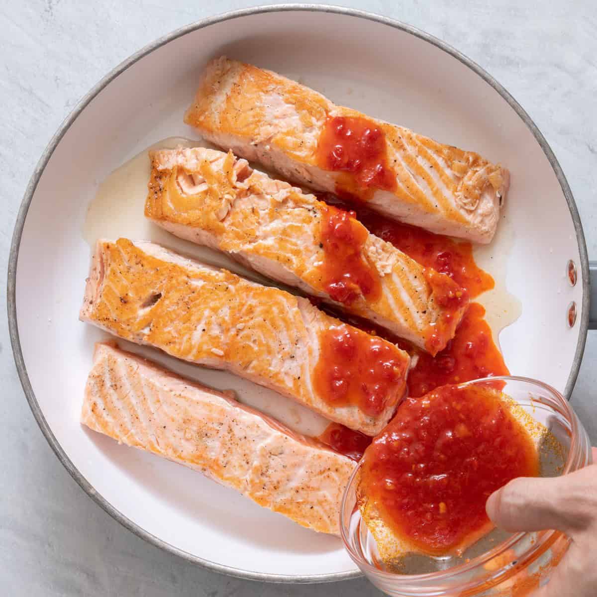 Salmon after searing in a pan with honey harissa sauce being poured on top.