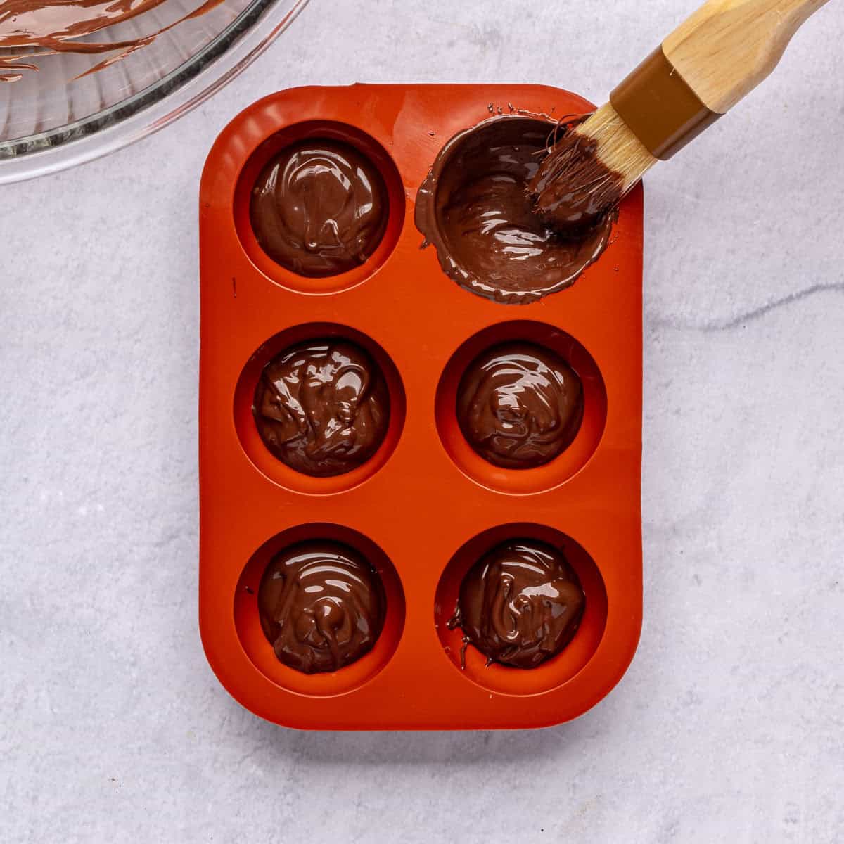 Brushing tempered chocolate into silicone molds with a tablespoon of chocolate in each one and the bowl of melted chocolate nearby.