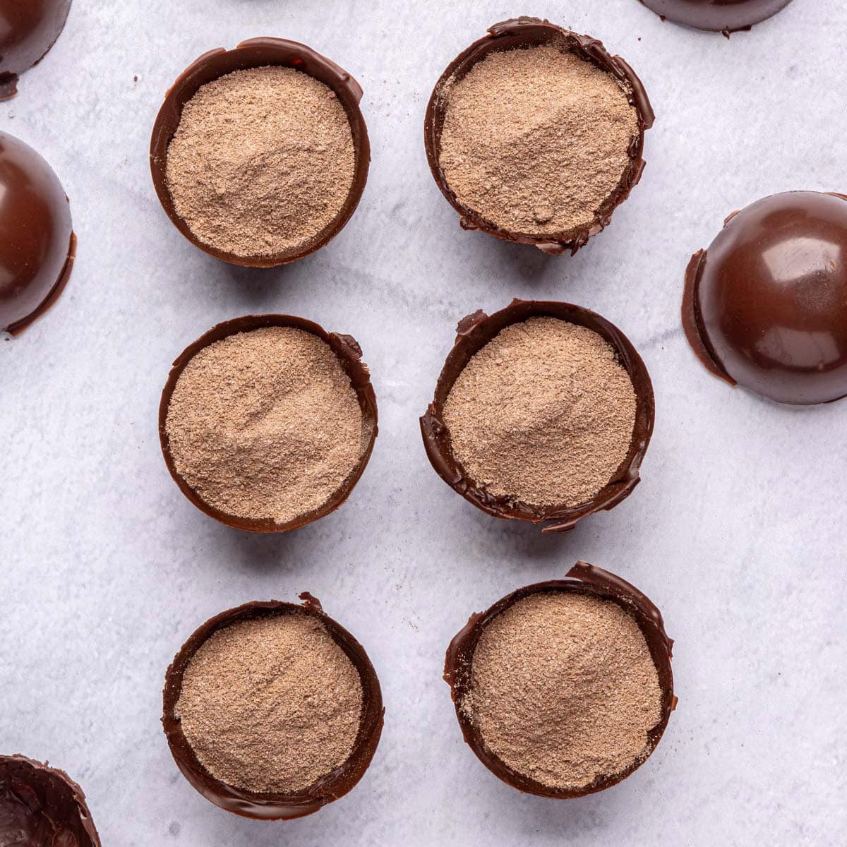 Cocoa mix filled to the brim inside 6 of the molds with the other shells (lids) nearby.
