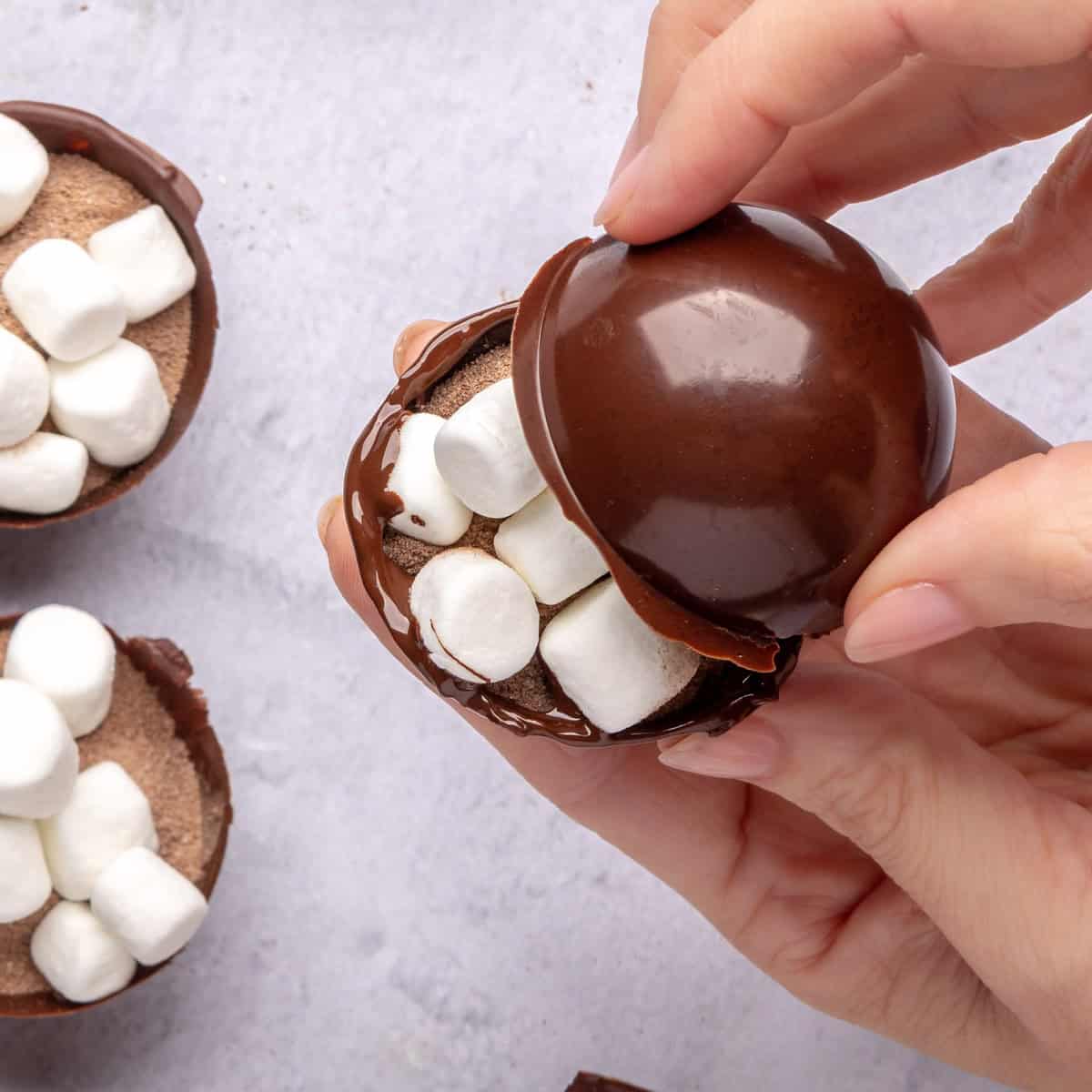 Hand holding filled chocolate shell with cocoa mix and mini marshmallows with top lid being added on top.