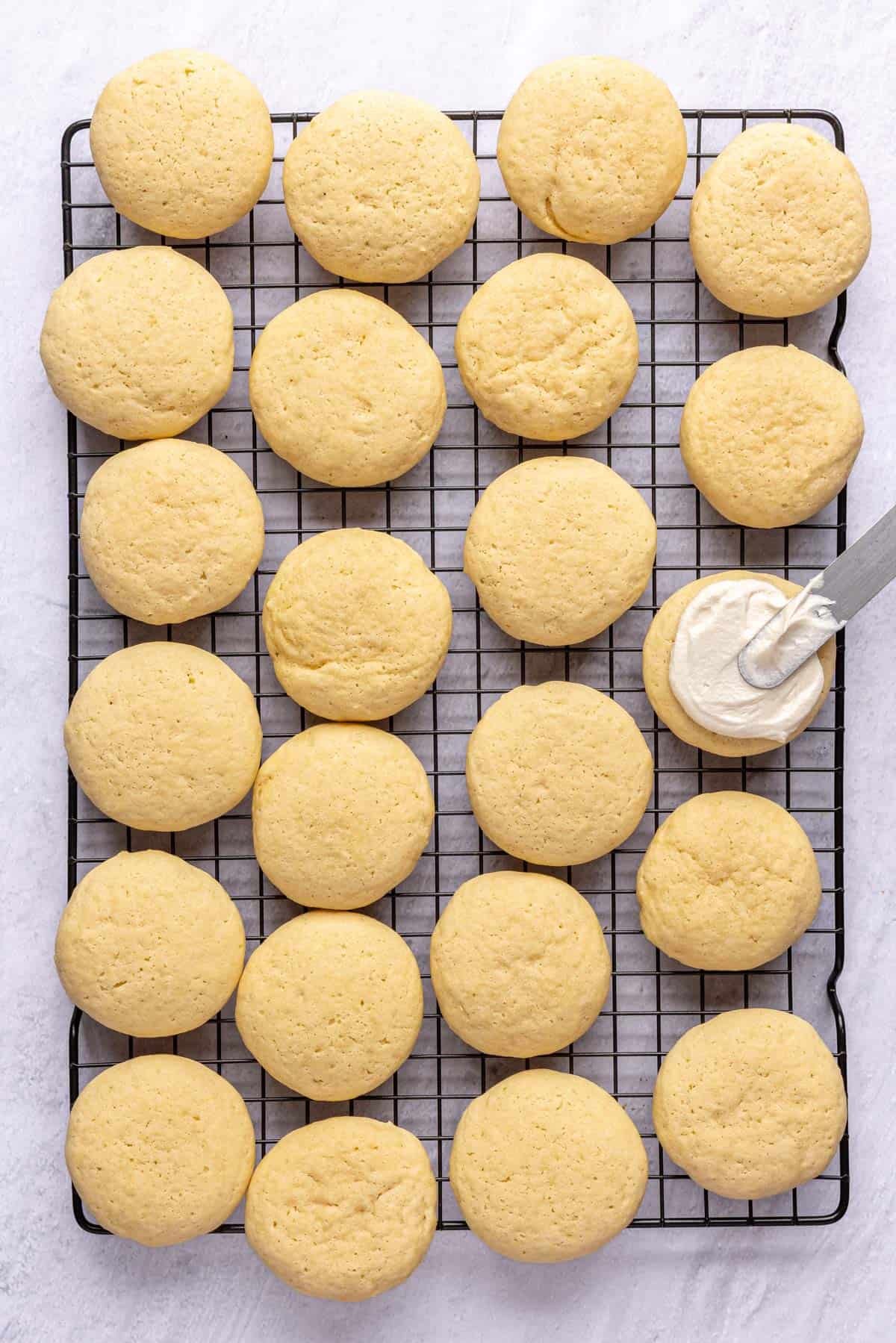 Baked Lofthouse sugar cookies on a wire rack after baking with frosting being spread onto cookie.