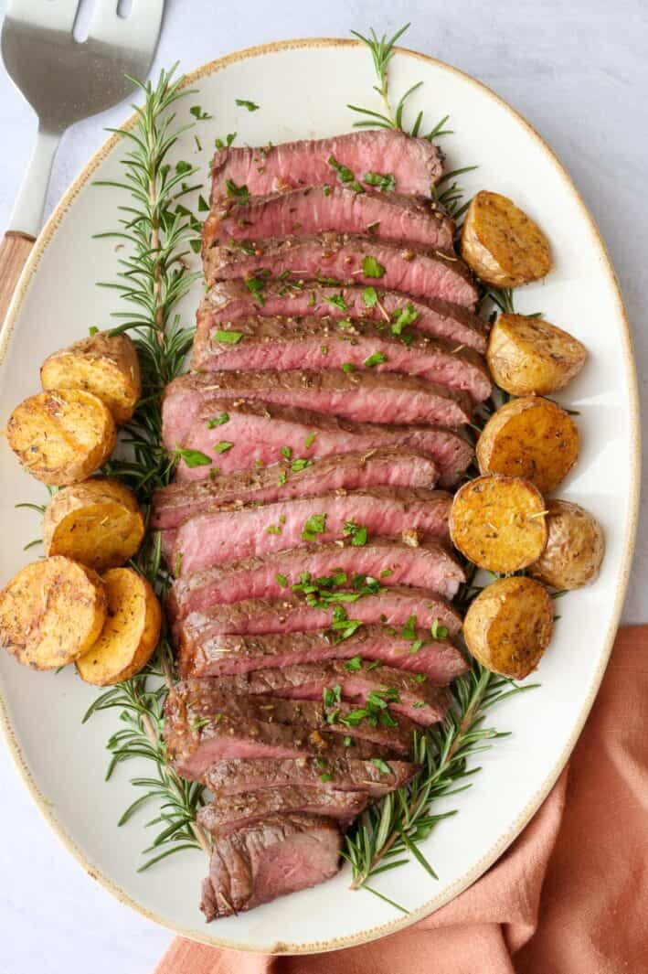 Thin slices of London broil on a platter with a few sprigs of fresh rosemary around and garnished with chopped parsley. Serving set resting nearby.