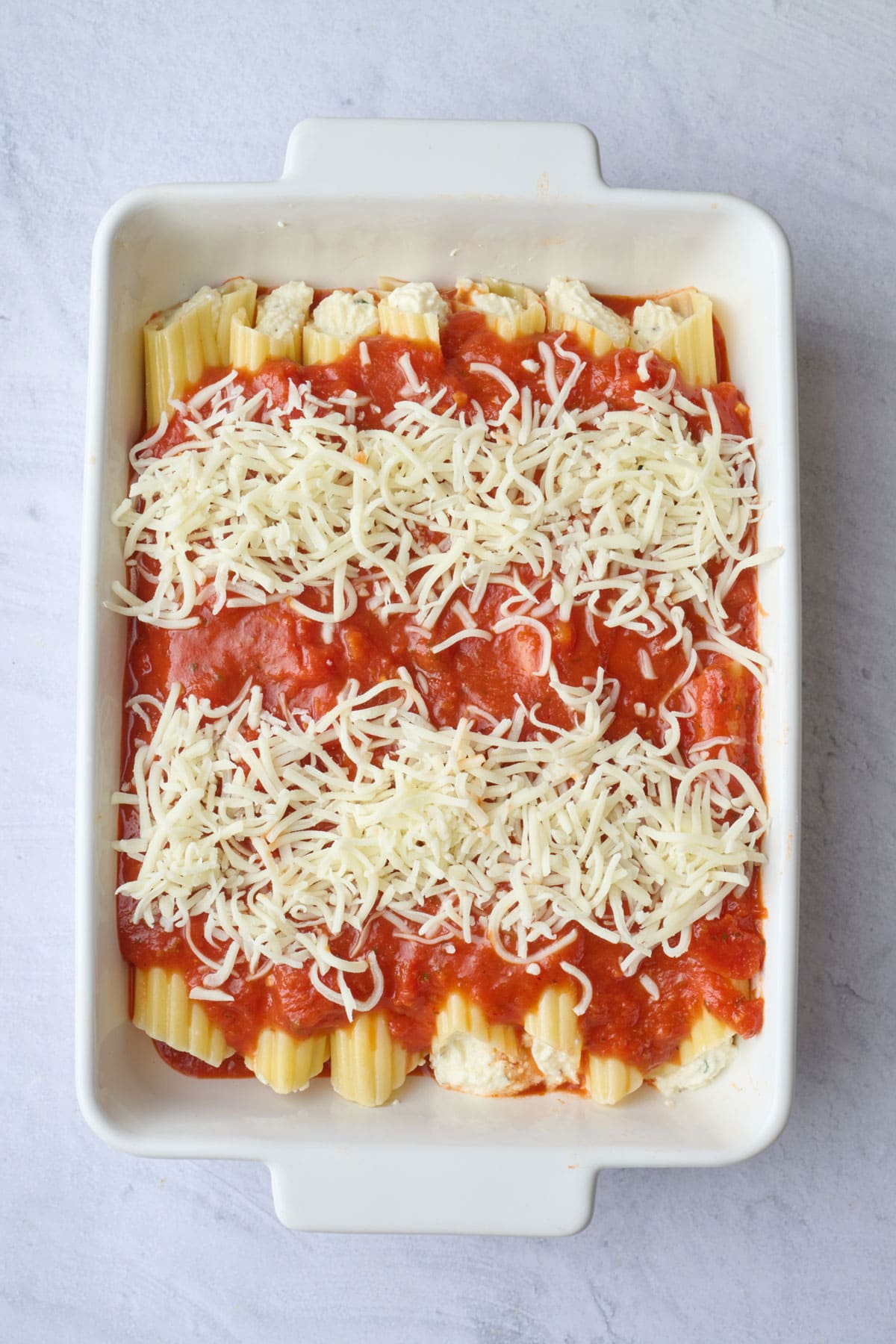 Marinara sauce spread into a baking dish with stuffed manicotti arranged into 2 width-wise rows, remaining sauce poured over the center of each row and mozzarella sprinkled on top before baking.