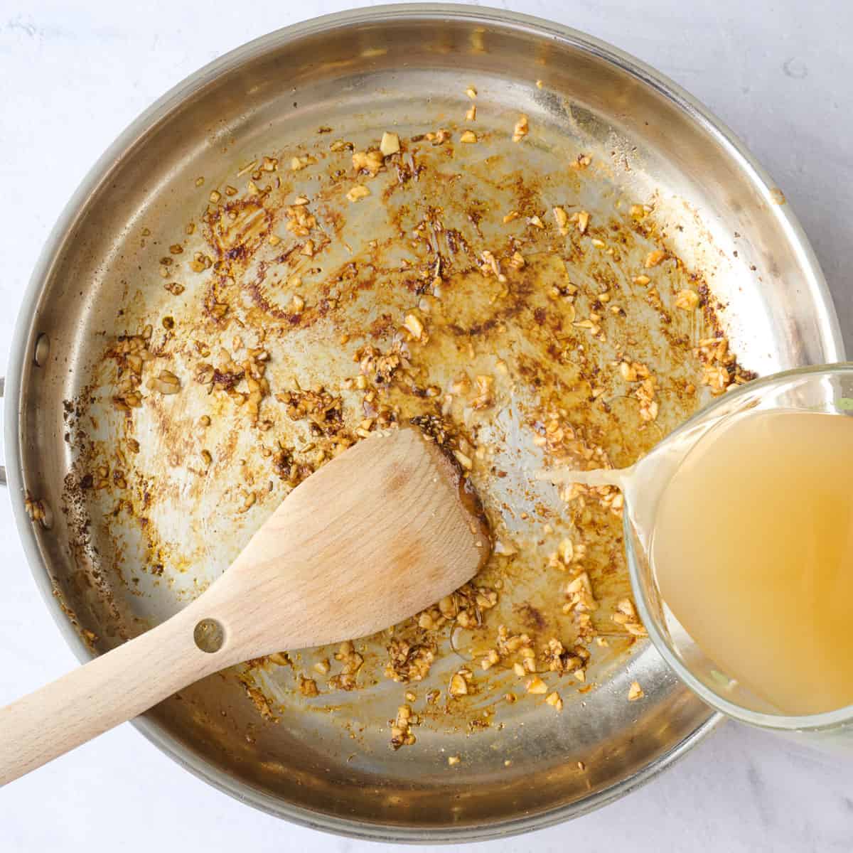 Broth being added to skillet while scraping up chicken bits and garlic from the bottom.