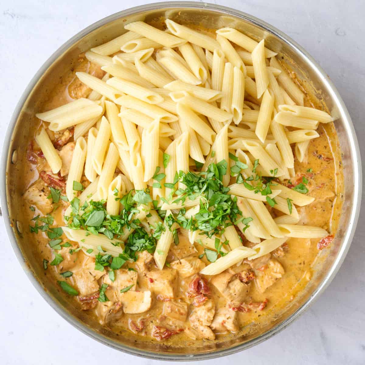 Marry me chicken pasta sauce in a skillet with cooked penne pasta and chopped basil added on top before tossing together.