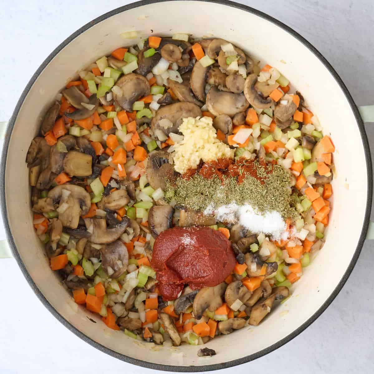 Cooked mirepoix with mushrooms in a pot with garlic, tomato paste, and seasonings added before combining for soup recipe.