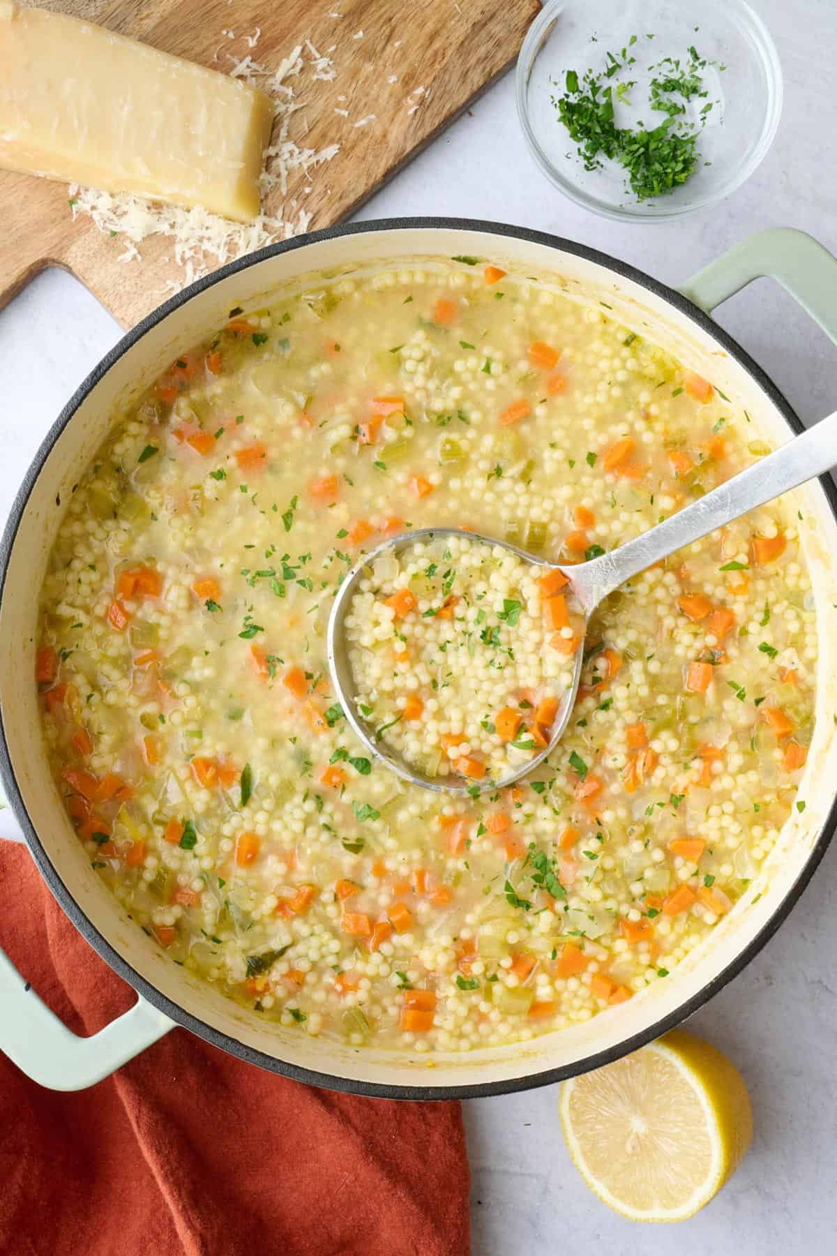Pastina soup recipe in the pot with a ladle lifting some up, small dish of parmesan and fresh parsley and lemon nearby.