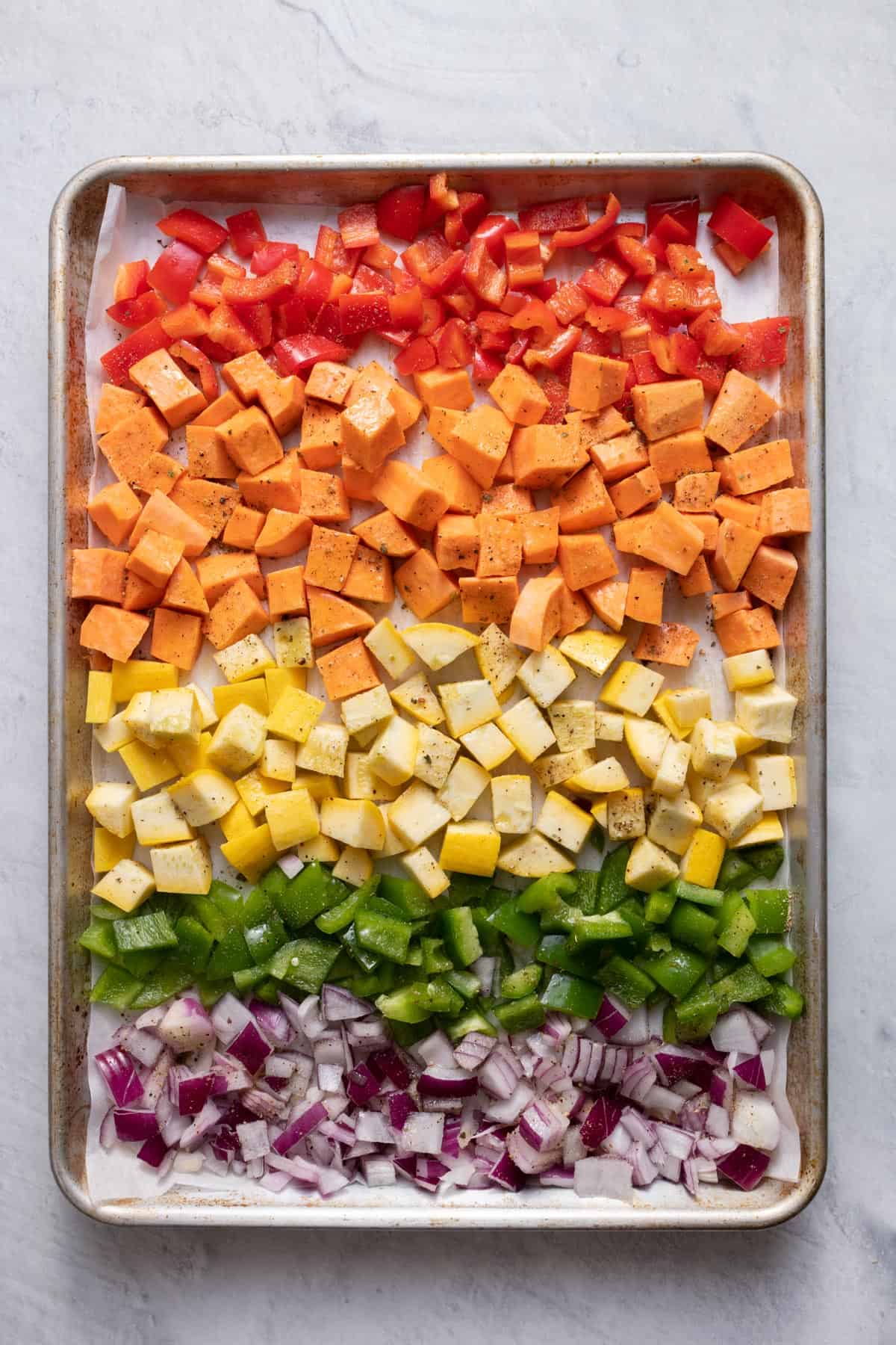 Diced red pepper, sweet potato, squash, green bell pepper and red onions on a sheet pan with seasonings on top before roasting.