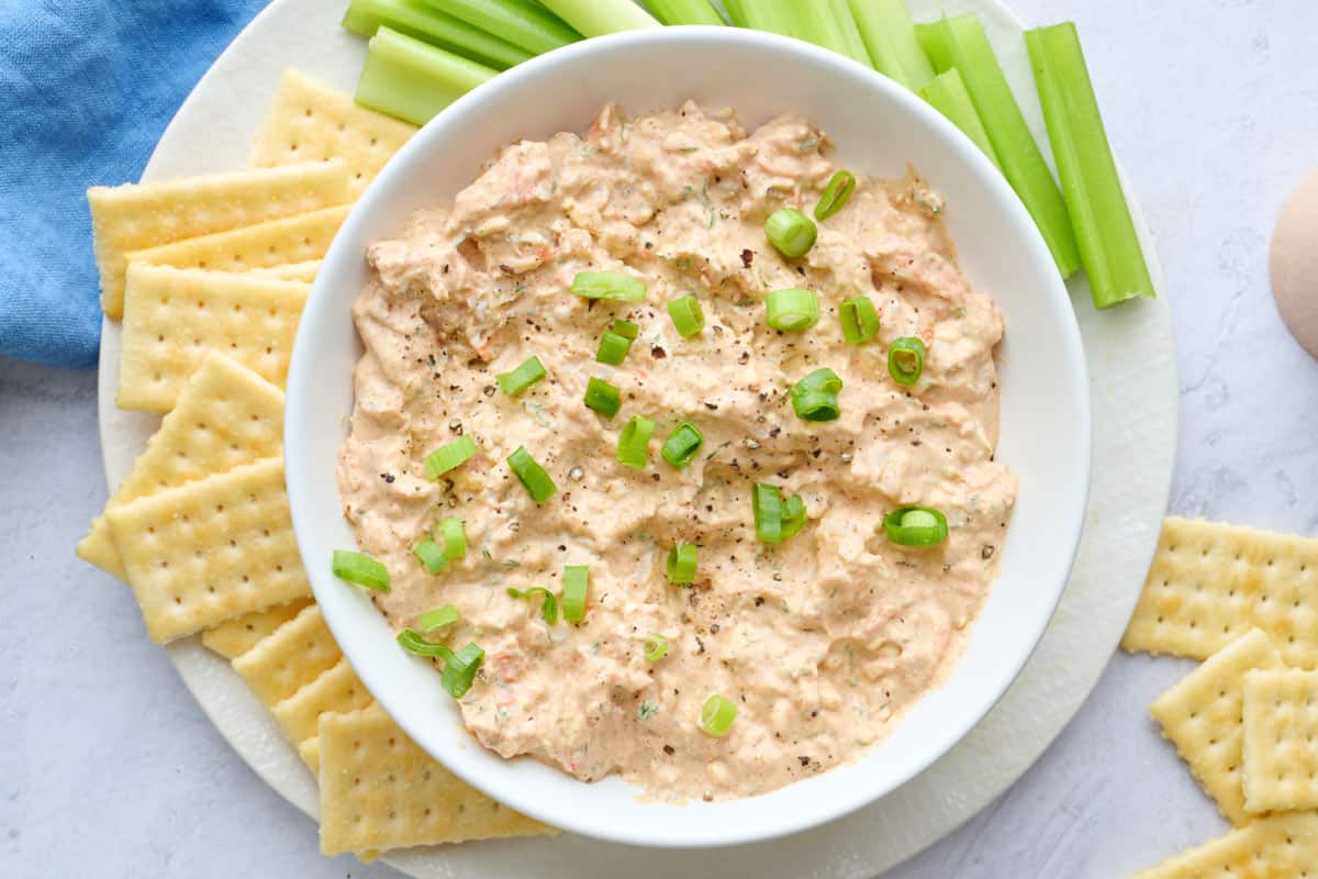 Cold shrimp dip appetizer in a serving bowl garnished with green onions and served on a plate with butter crackers and celery sticks.