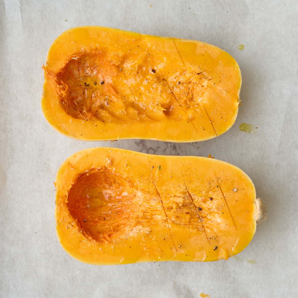 Two halves of a butternut squash with seeds scooped out on parchment paper after coating with oil and spices before roasting.