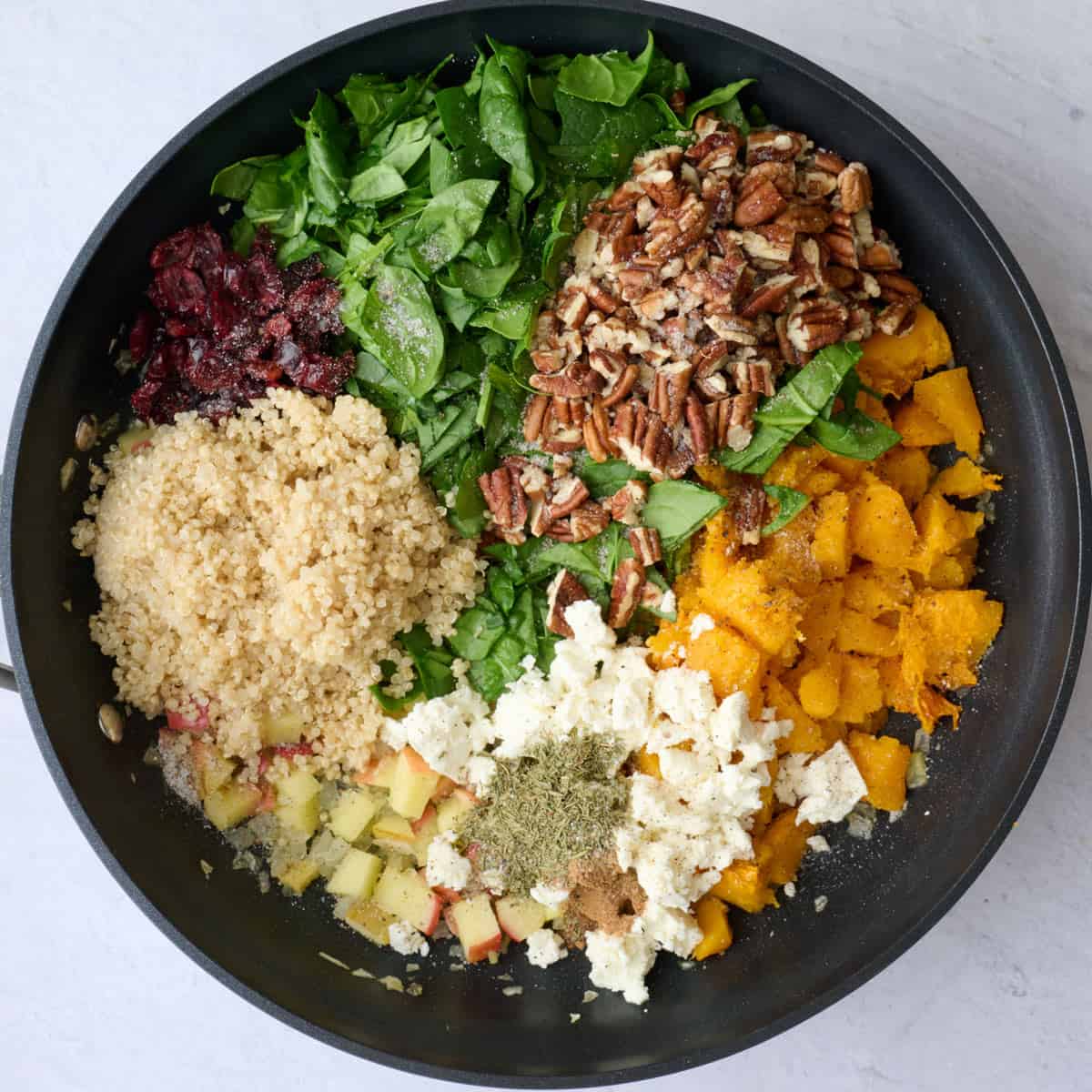 Cooked quinoa, dried cranberries, chopped spinach, pecans, feta cheese, seasonings, and butternut squash in a skillet with the apple onion mixture before tossing together.