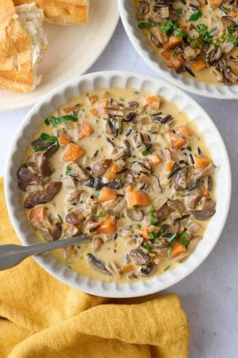 Two bowls of mushroom soup with wild rice with focus on one, a few sliced mushrooms pulled to the top with a spoon dipped inside, torn bread nearby.