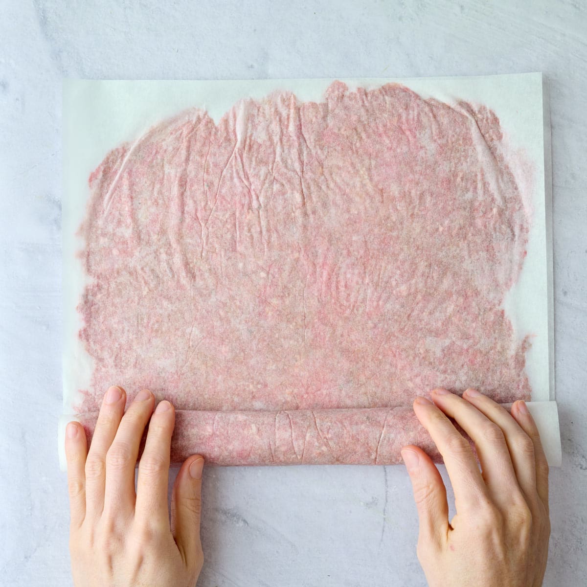 Hands rolling the thin parchment covered sheet of ground beef mixture into a tight log for baking.