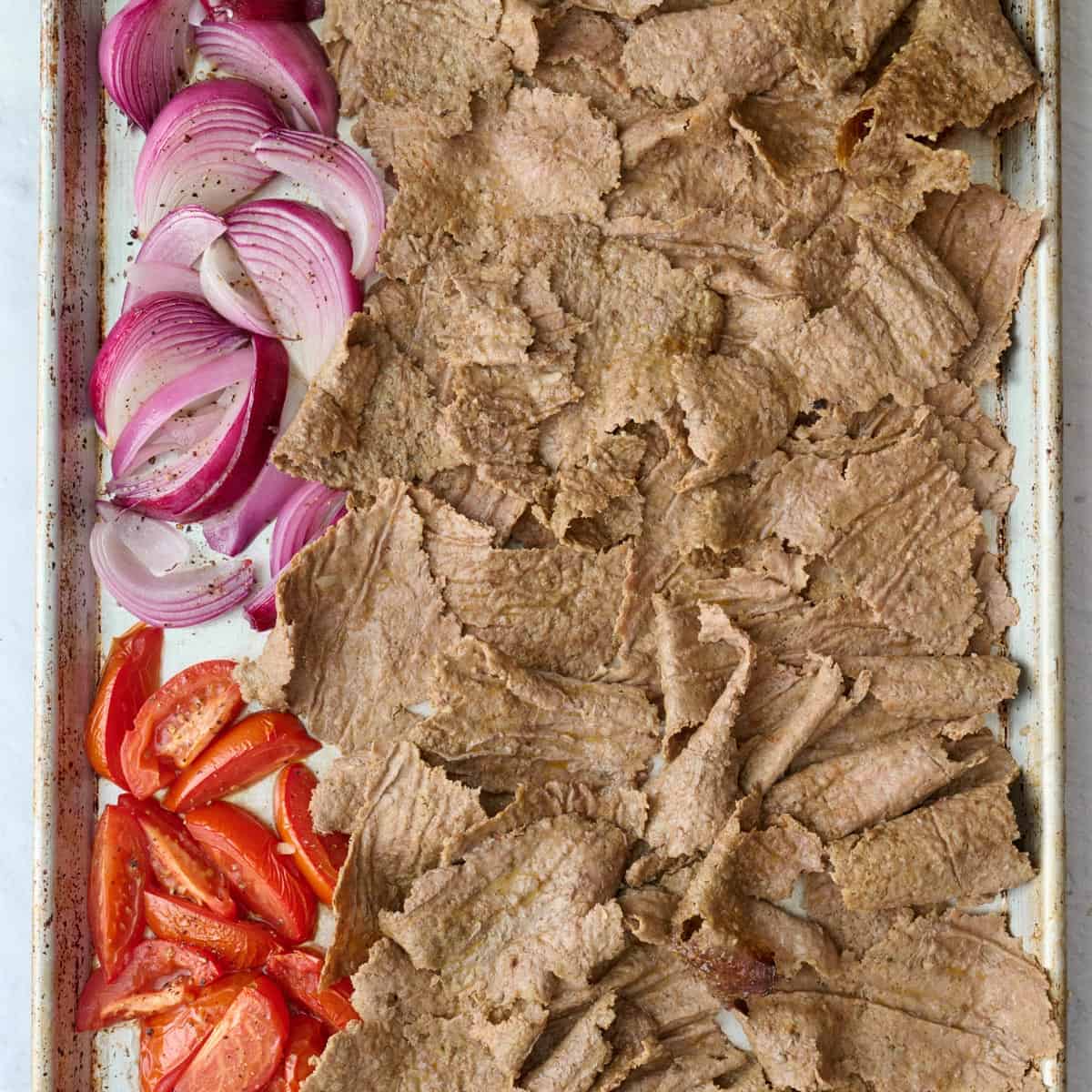 Cooked doner kebab meat after cooked and broken into pieces on a sheet pan with roasted onions and tomatoes before broiling.