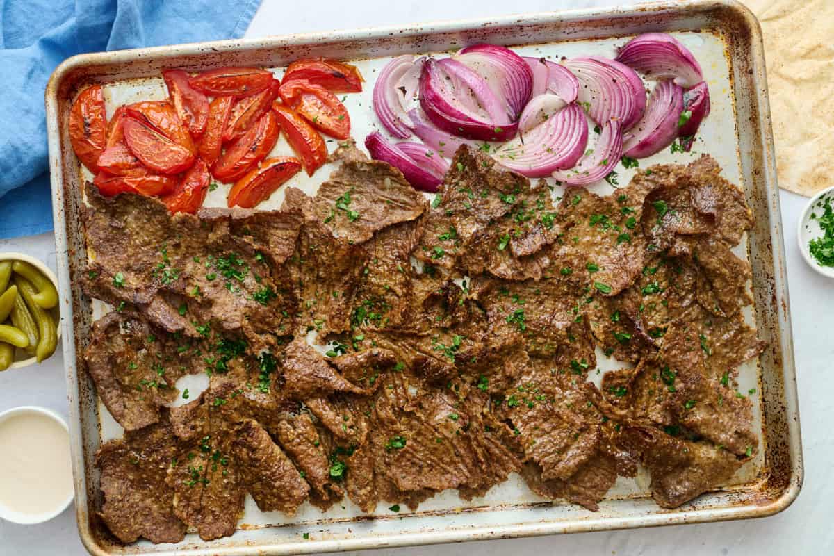 Beef doner kebab meat with roasted tomato and onions on a sheet pan for serving with pita, pickles and tahini sauce nearby.