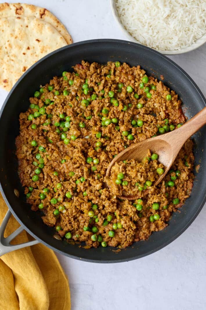 Keema curry with seasoned ground beef and peas in a skillet with serving spoon dipped inside and a bowl of cooked rice and naan nearby.
