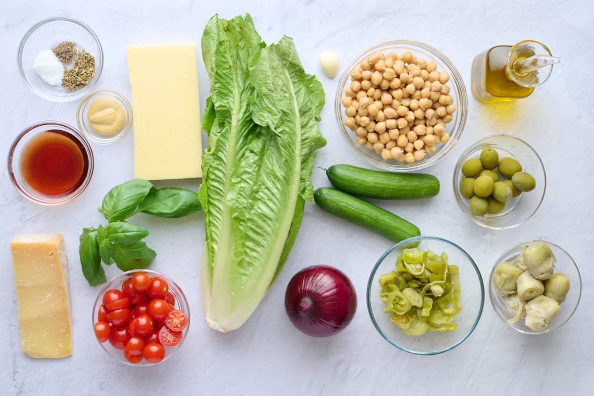 Ingredients for salad recipe: Italian seasoning, salt and pepper, red wine vinegar, Dijon mustard, parmesan, fresh basil, tomatoes, mozzarella cheese, romaine lettuce, garlic, chickpeas, cucumbers, red onion, pepperoncini, chopped artichoke hearts, olives and oil.