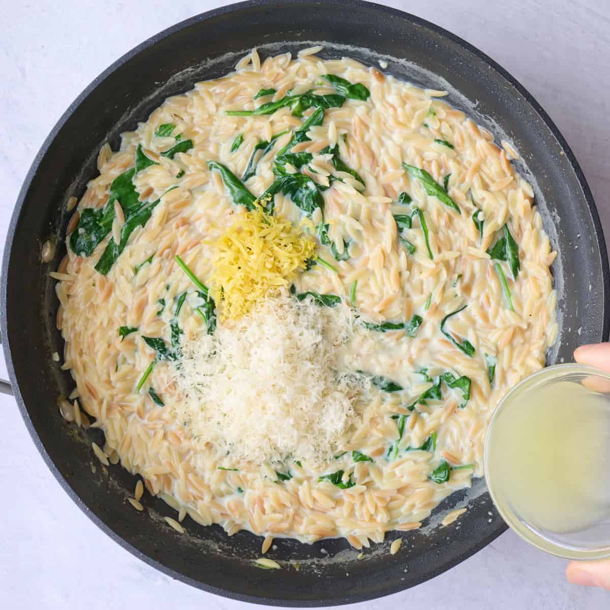 Creamy spinach orzo in a pan with lemon and grated parmesan added before combining.