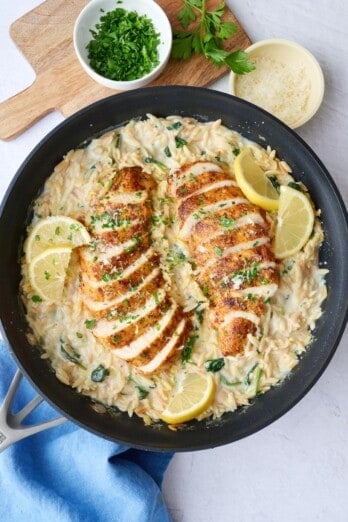 Lemon chicken orzo in the pan with chicken breast sliced and fanned out on top, garnished with parsley and lemon wedges.