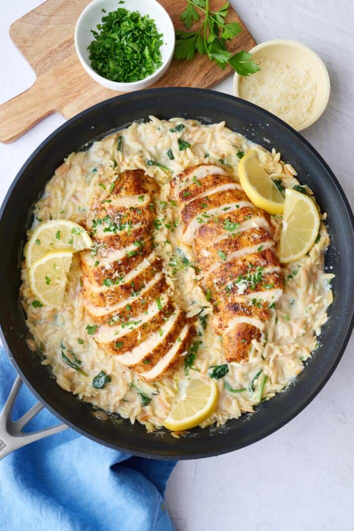 Lemon chicken orzo in the pan with chicken breast sliced and fanned out on top, garnished with parsley and lemon wedges.