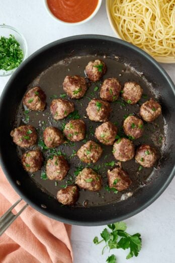 Easy beef meatball recipe in a skillet with a side of cooked pasta and marinara sauce nearby.