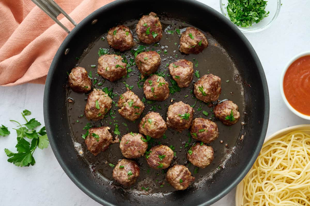 Best ground beef meatball recipe in a skillet with a side of cooked pasta and marinara sauce nearby.