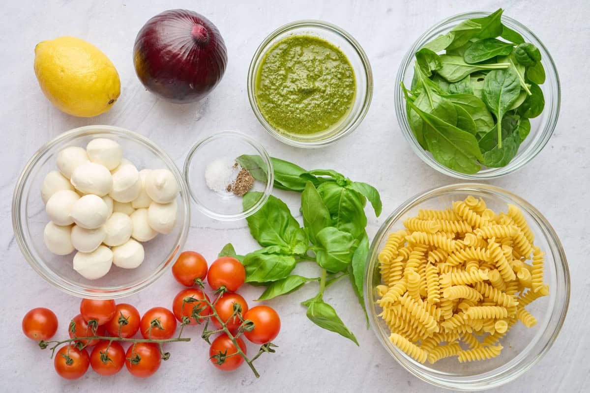 Ingredients for salad recipe: lemon, red onion, mozzarella pearls, cherry tomatoes, salt and pepper, pesto, basil, spinach, and fusilli.