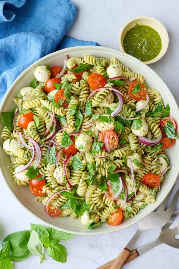Pesto salad in a serving bowl garnished with fresh chopped basil, serving set dipped inside, small dish of pesto nearby.