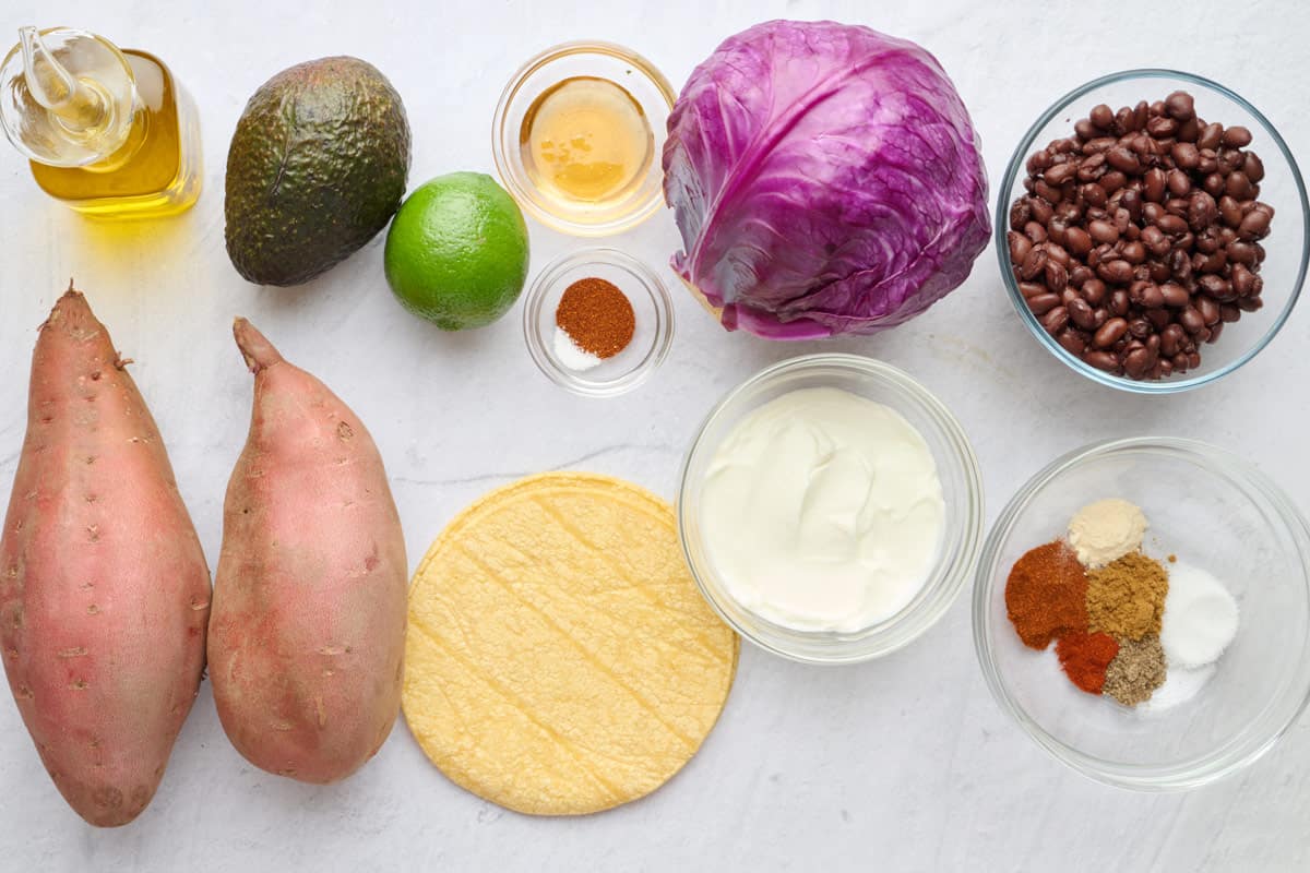Ingredients for taco recipe: sweet potatoes, oil, avocado, lime, honey, chili powder and salt, corn tortillas, red cabbage, yogurt, black beans, and seasonings.