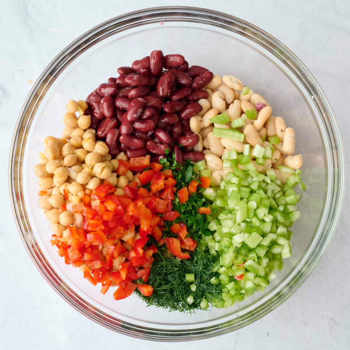 Chickpeas, kidney beans, white beans, chopped celery, red pepper and fresh herbs segmented in a bowl before tossing together.