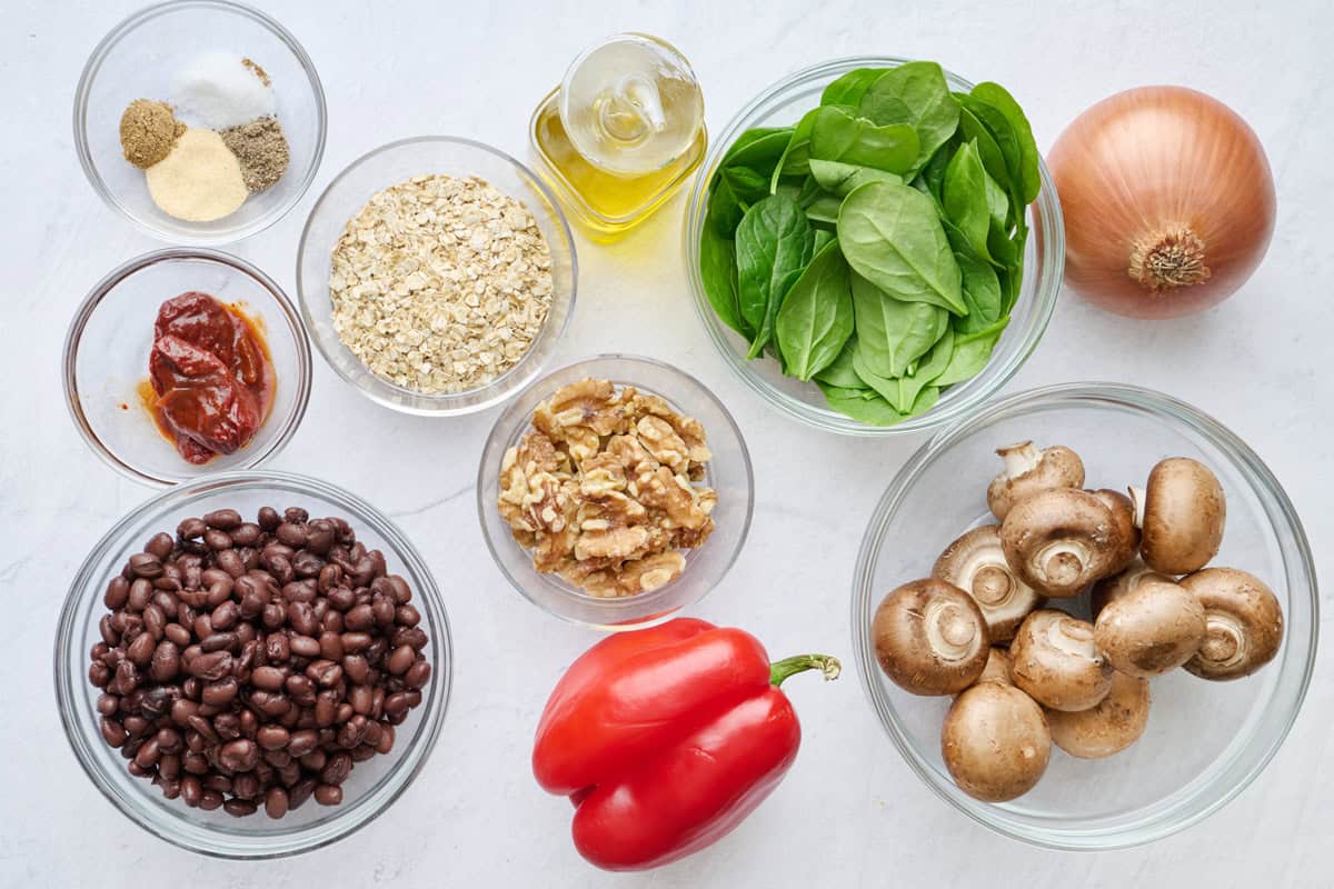 Ingredients for veggie burger recipe: seasonings, chipotle peppers in adobe sauce, black beans, red bell pepper, walnuts, oats, oil, spinach, mushrooms, and onion.