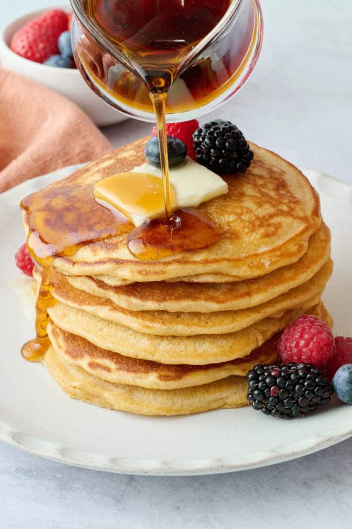 Homemade whole wheat pancakes on a plate with maple syrup being poured on top.