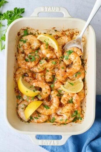 Garlic butter baked shrimp recipe in a baking dish garnished with parsley and lemon wedges with a spoon dipped in.