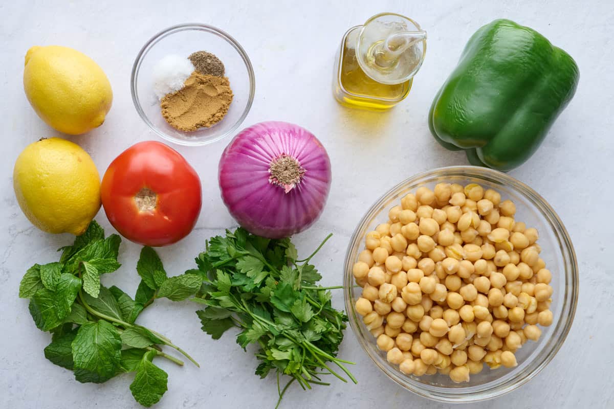 Ingredients for bean salad recipe: lemon, tomato, red onion, fresh parsley and mint, spices, oil, chickpeas, and bell pepper.