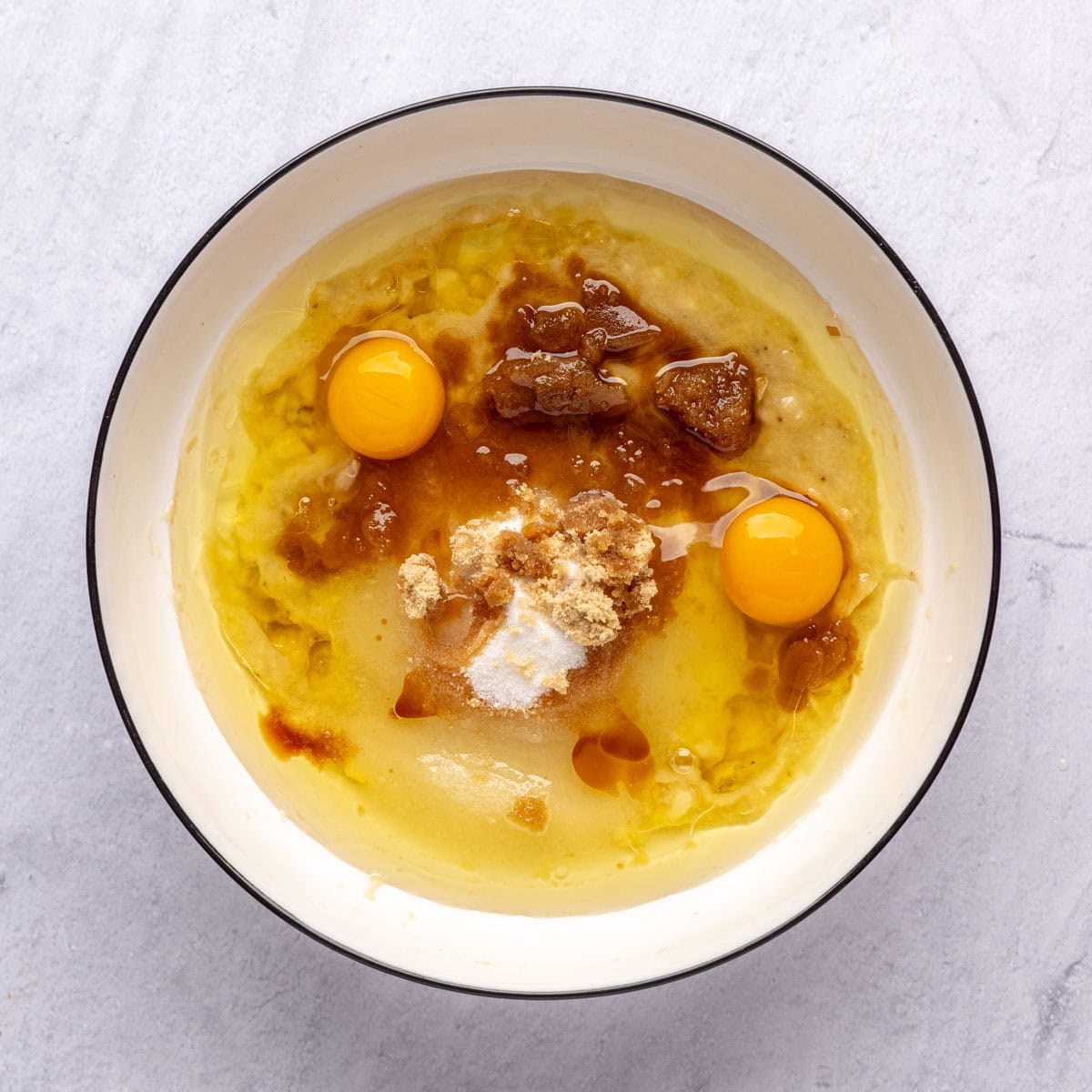 Mashed bananas in a bowl with oil, sugars, eggs and vanilla added before combining.