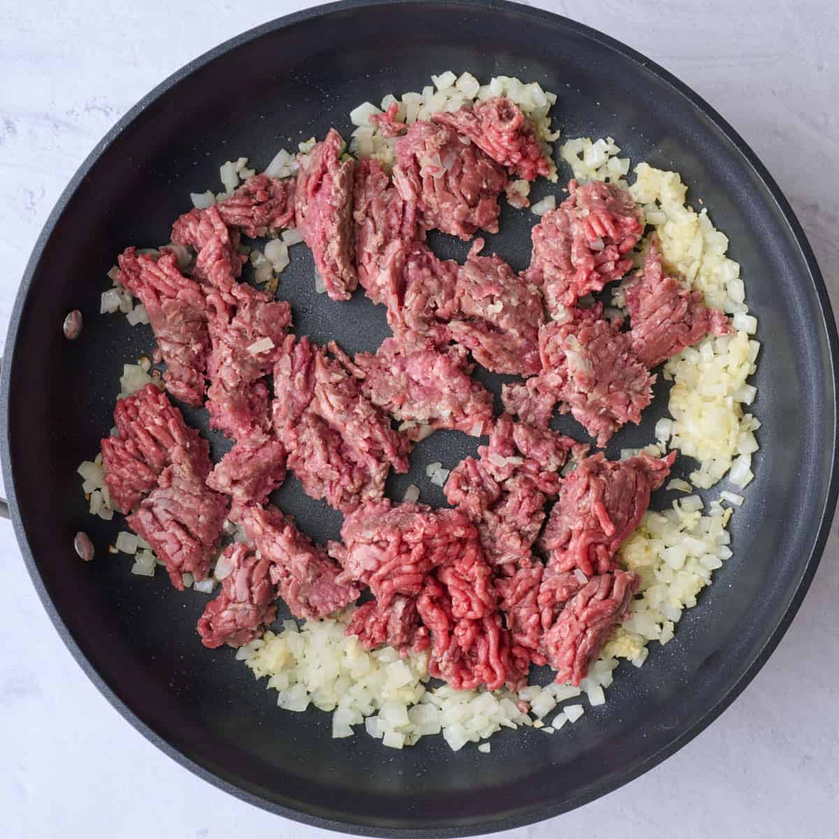 Cooked onion and garlic in a skillet with ground beef added, broken into pieces, before browning.