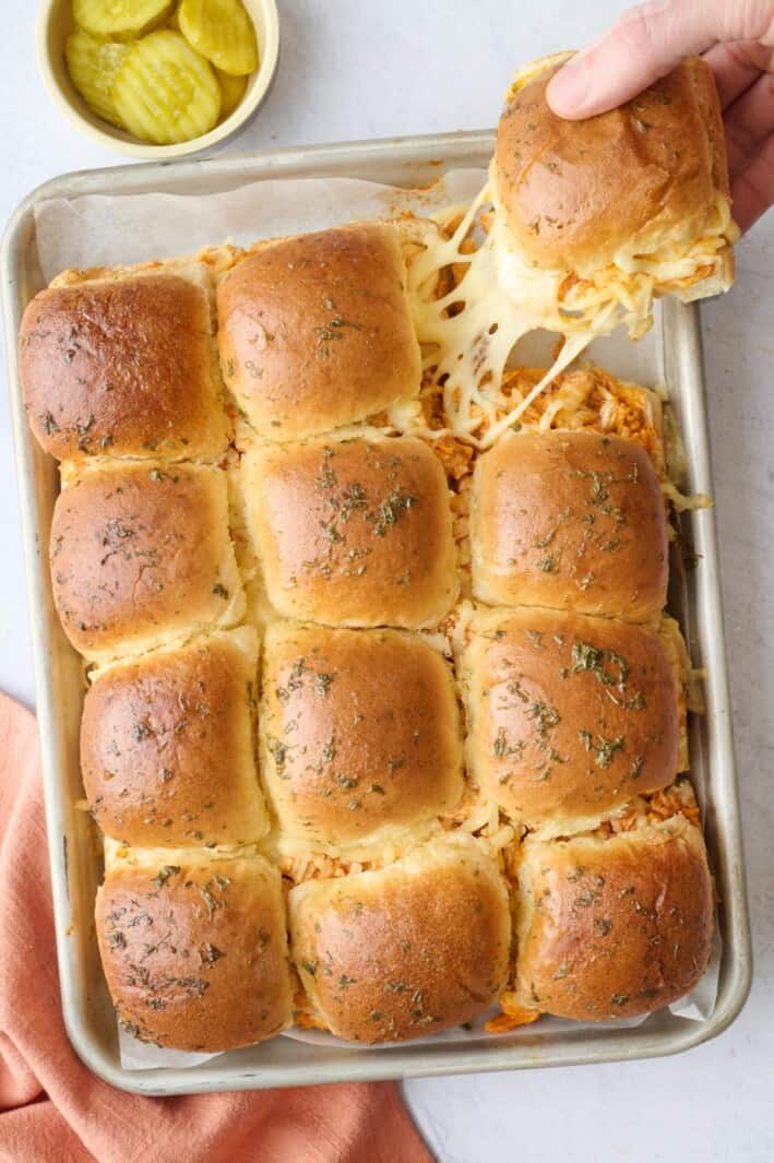 Easy buffalo chicken sliders on a baking sheet with one piece being grabbed with a cheesy pull.
