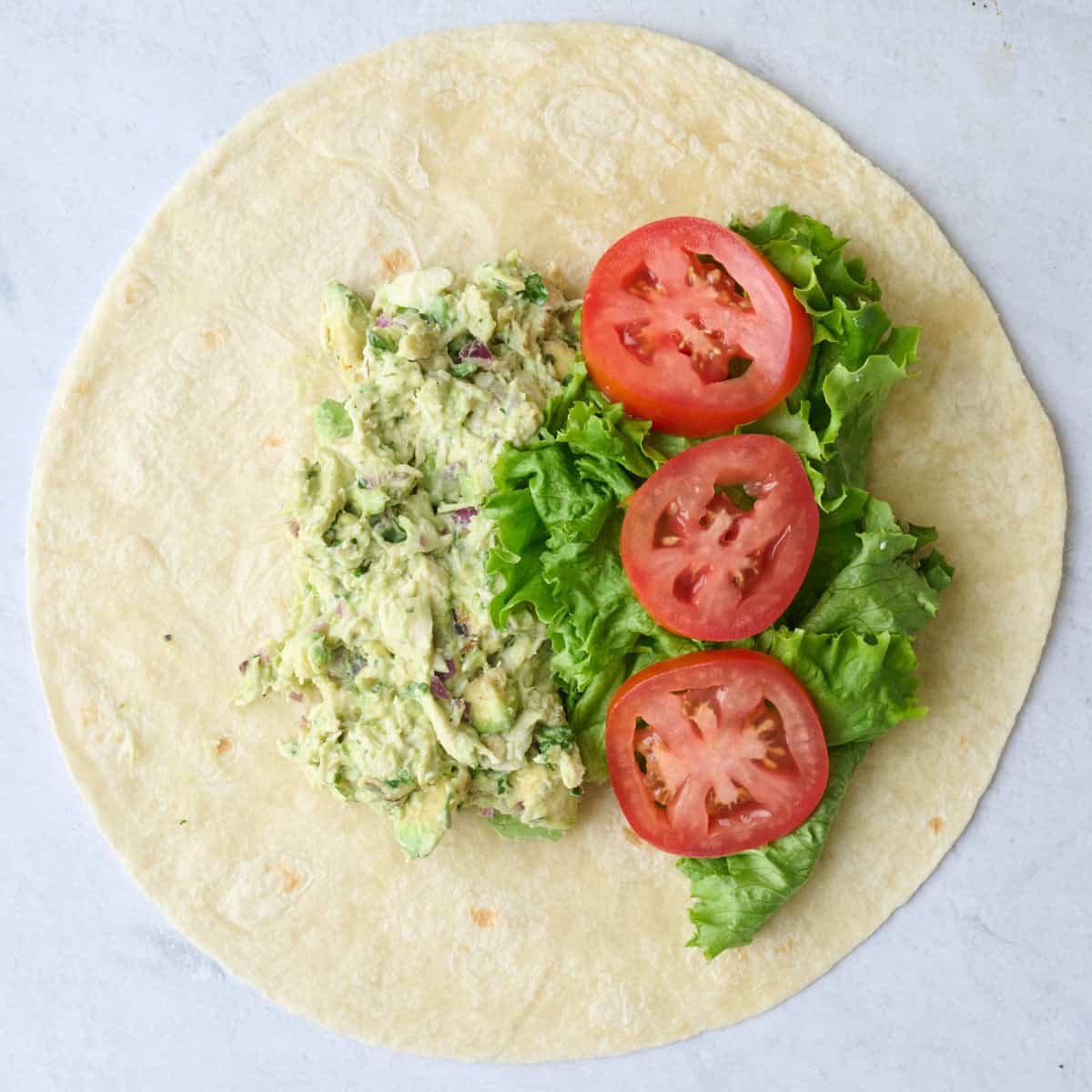Prepared chicken avocado salad mixture added to a tortilla shell with lettuce and tomato slices before wrapping.
