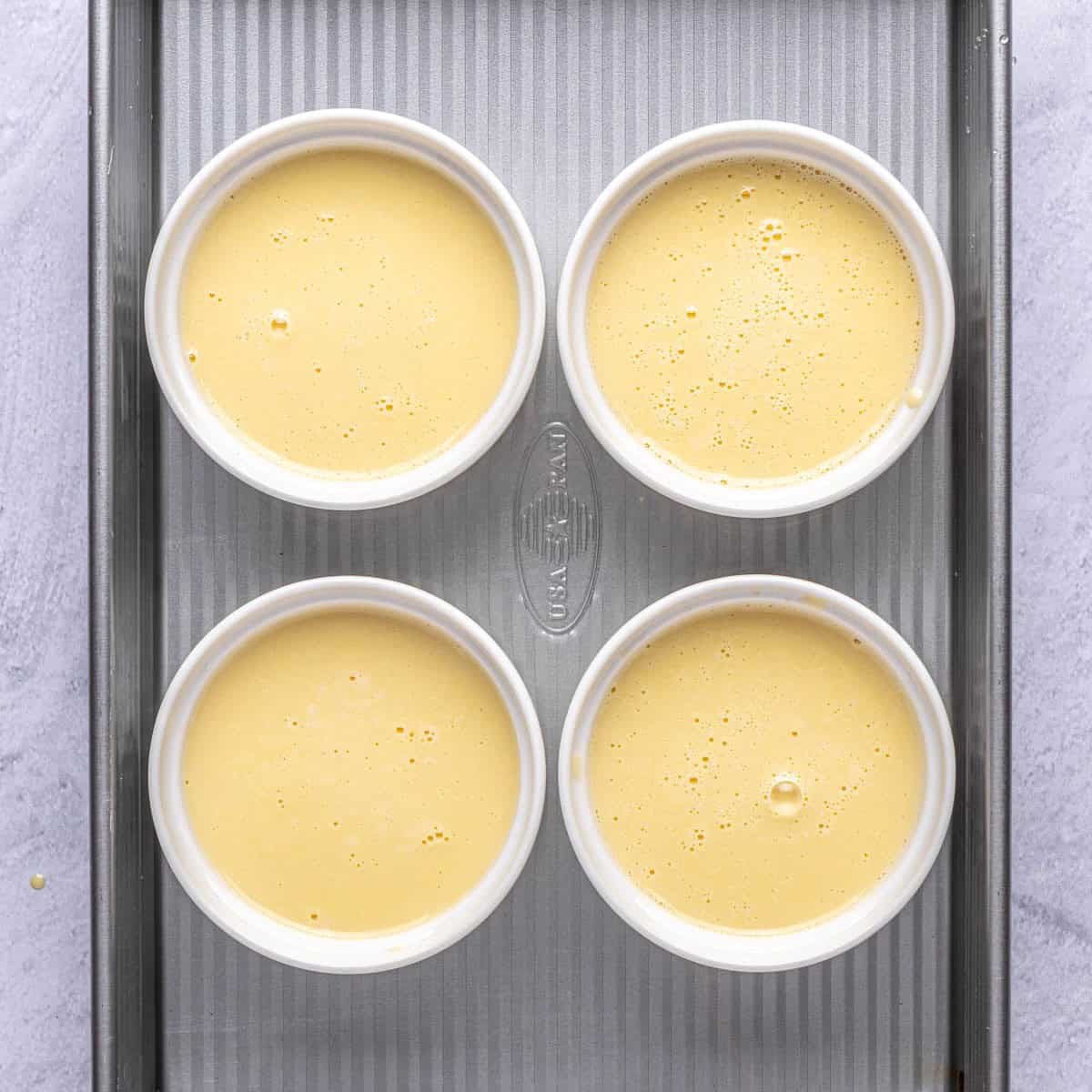 Vanilla custard divided into 4 ramekins setting in a baking dish with hot water halfway up before baking.
