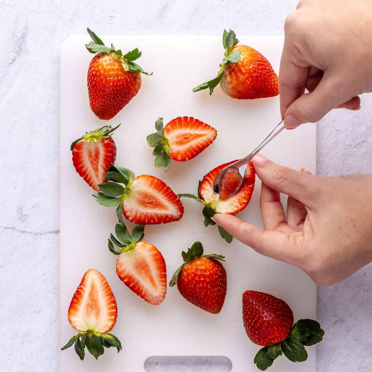 A few strawberries on a small cutting board sliced in half lengthwise with a small spoon being used to hollow out.