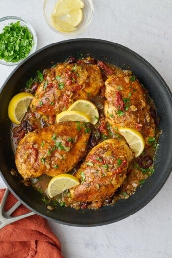 Best Mediterranean Chicken breast in a skillet with sauce with a little extra parsley on top, a few lemon wedges nearby.