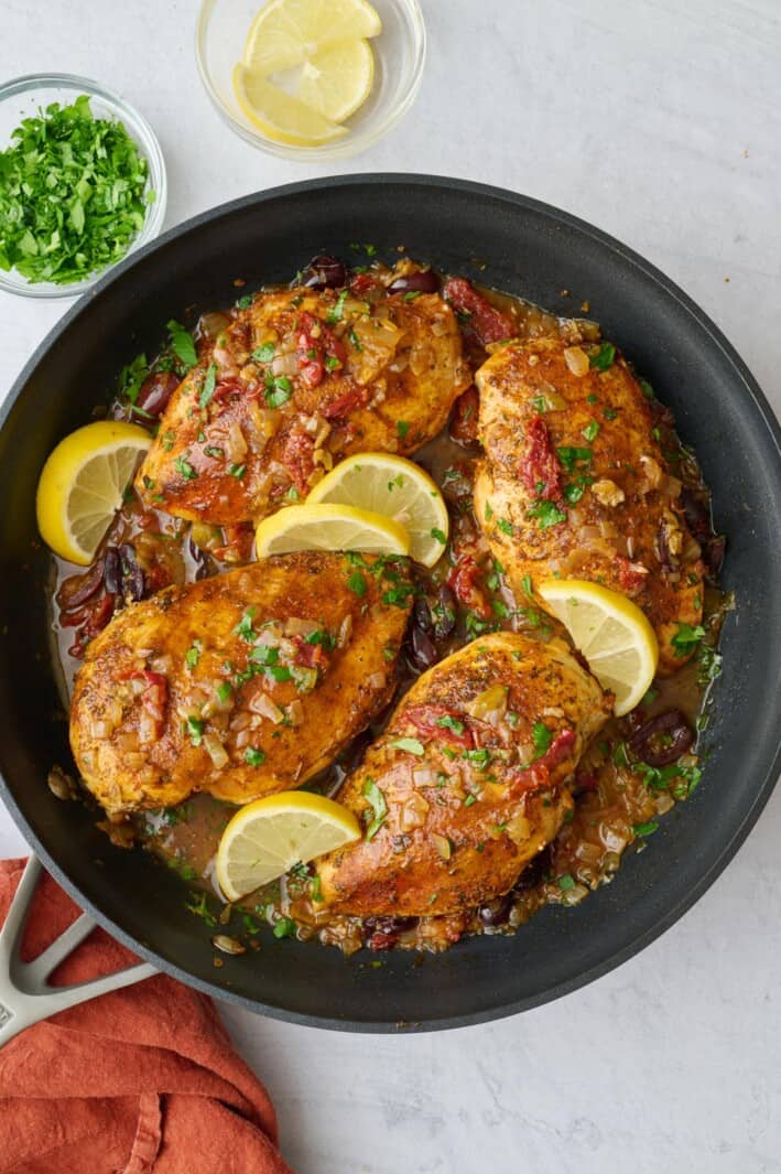 Best Mediterranean Chicken breast in a skillet with sauce with a little extra parsley on top, a few lemon wedges nearby.