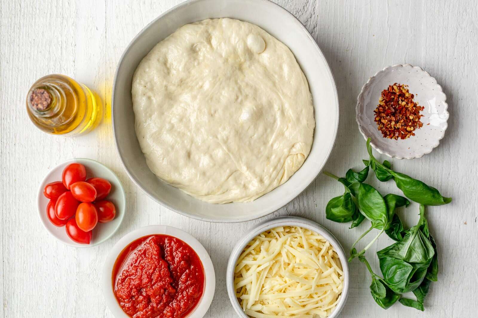Ingredients for pizza recipe: oil, tomatoes, tomato sauce, pizza dough, shredded cheese, basil, and crushed red pepper flakes.