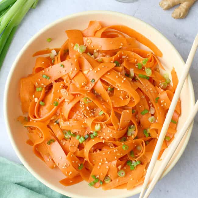 Carrot salad in a serving bowl garnished with sesame seeds, chopsticks dipped inside, small dish of extra sesame seeds nearby