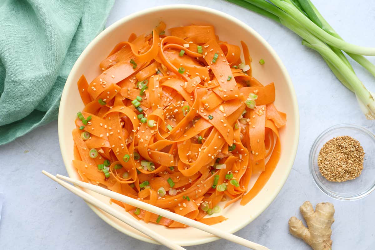 Carrot salad in a serving bowl garnished with sesame seeds, chopsticks dipped inside, small dish of extra sesame seeds nearby