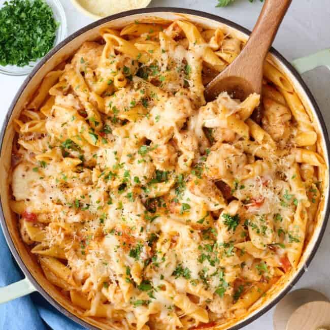 Chicken Parmesan Pasta in a pot with a wooden spoon, with ingredients for the recipe surrounding.