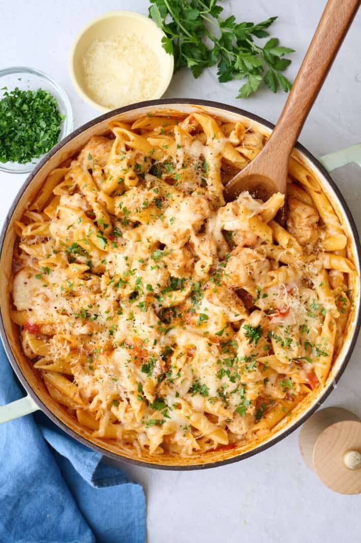 Chicken Parmesan Pasta in a pot with a wooden spoon, with ingredients for the recipe surrounding.