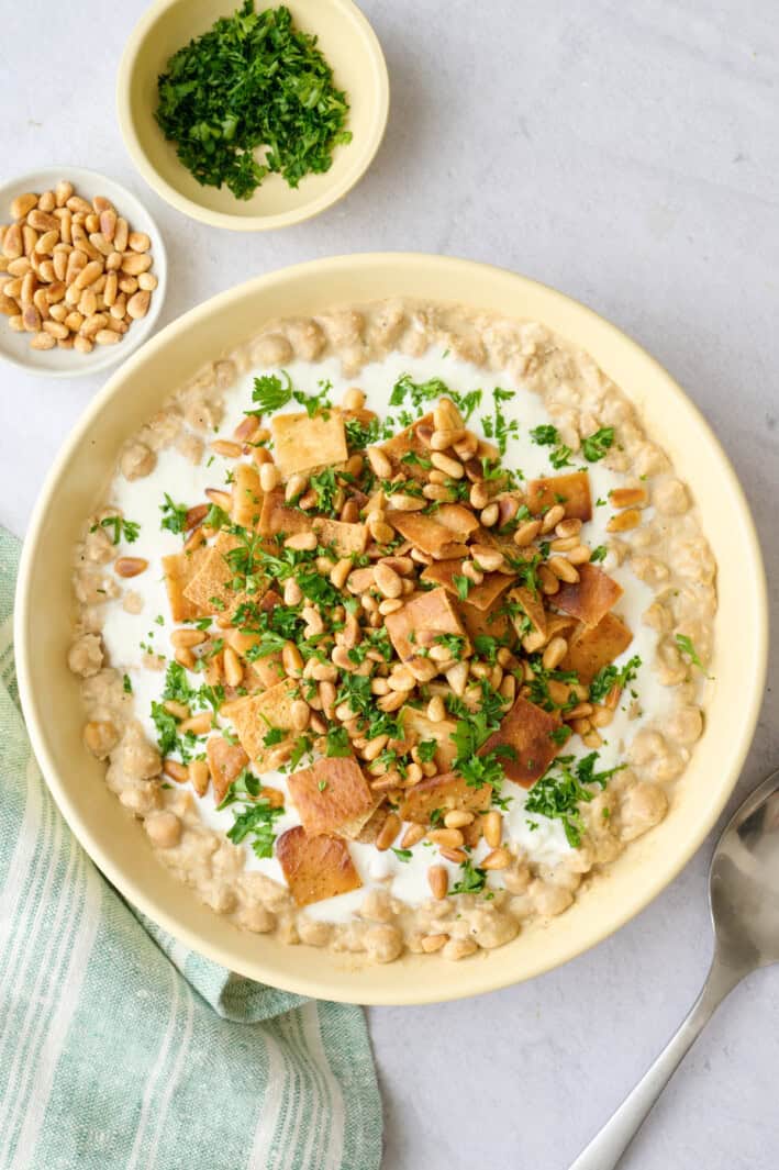 Fatteh hummus recipe layer in a bowl with mashed chickpeas, yogurt sauce, toasted pita, pine nuts and parsley.